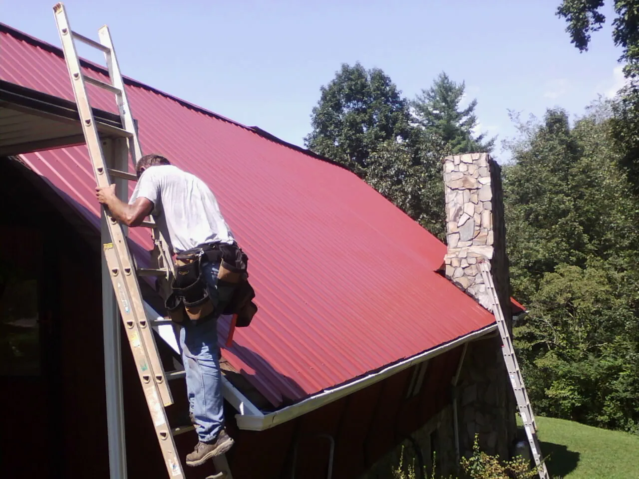 Roofer on a ladder beside a red metal roof and stone chimney — Soffit & Fascia Repair by Roofing Brothers in Lenhartsville